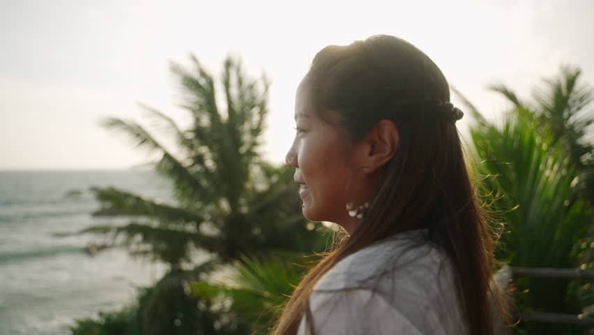 Asian woman in white dress looking at sea. Ethnic young brunette female watching sunset on authentic balcony on tropical location. Tanned delighted biracial girl admiring ocean view at dusk.