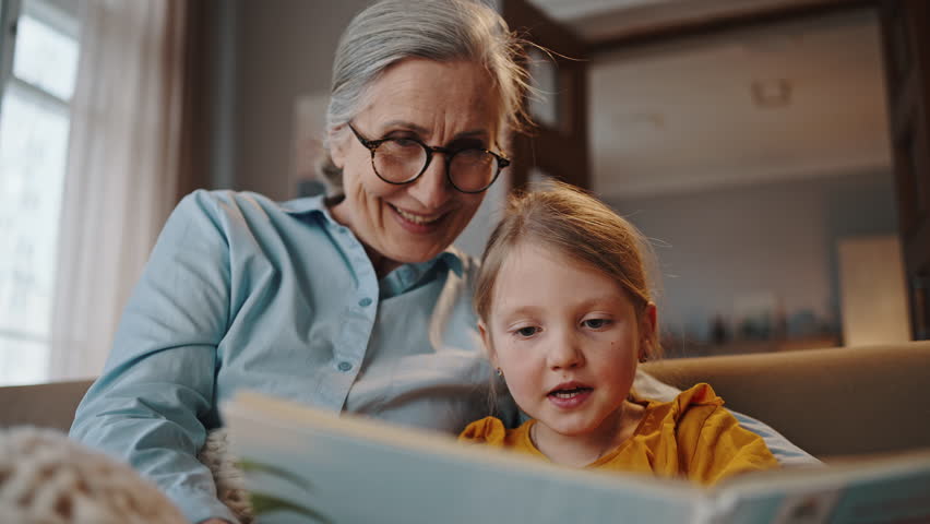 Close up of an adorable Caucasian redhead little child girl, reading book together with her elderly loving grandmother in the home living room. Happy family relationships concept. Three generations