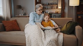 Adorable little child girl sits near her loving grandmother under cozy blanket on the sofa at home living room, smiles and feels proud to read a fairy tale. Grandma teaches her granddaughter alphabet - Powered by Shutterstock - Get 15% off with code: PIKWIZARD15