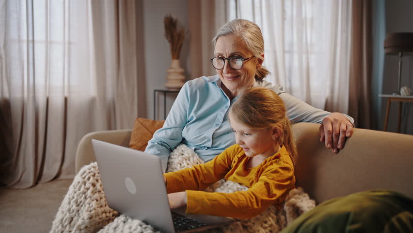 Beautiful Caucasian gray haired senior woman, a caring loving grandmother and granddaughter using laptop, browsing web, doing online homework while sitting together on sofa at cozy home interior Royalty-Free Stock Footage #1102569005