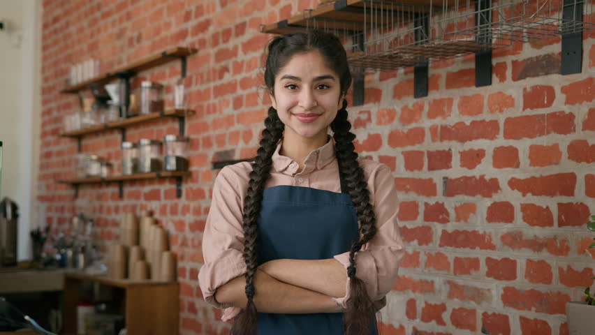 Portrait smiling indian woman waitress barista in apron posing in cafe cafeteria with crossed arms confident arabian girl female small business cafe owner looking at camera in restaurant cafeteria bar Royalty-Free Stock Footage #1102571703