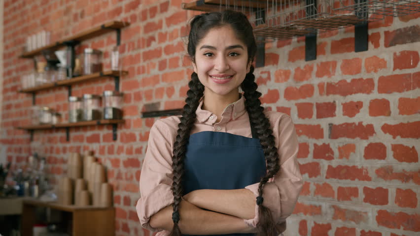 Portrait smiling indian woman waitress barista in apron posing in cafe cafeteria with crossed arms confident arabian girl female small business cafe owner looking at camera in restaurant cafeteria bar