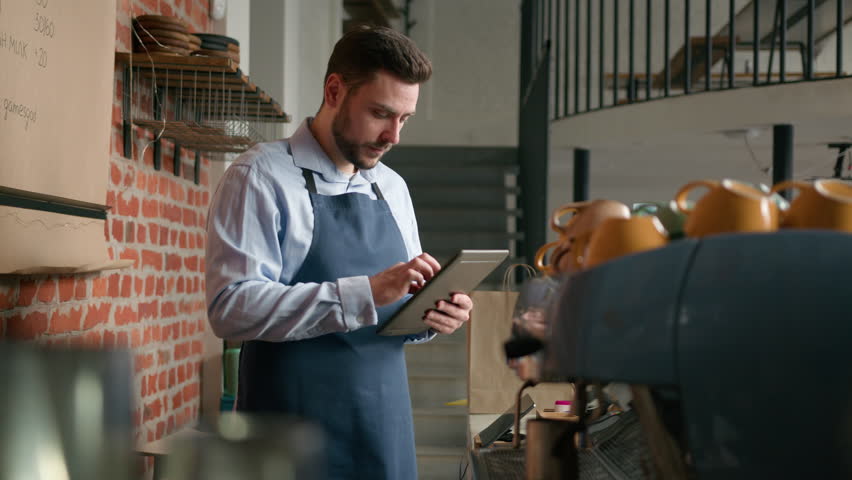 Caucasian man waiter barista cafe owner businessman using digital tablet to manage small business male manager restaurant worker in apron remote making order online standing behind cafeteria counter | Shutterstock HD Video #1102571705