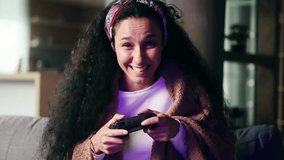 Camera view of excited young hispanic woman gamer sitting on a couch and playing in video games on console at home Happy player curly female controlling joystick for online competition indoors alone
