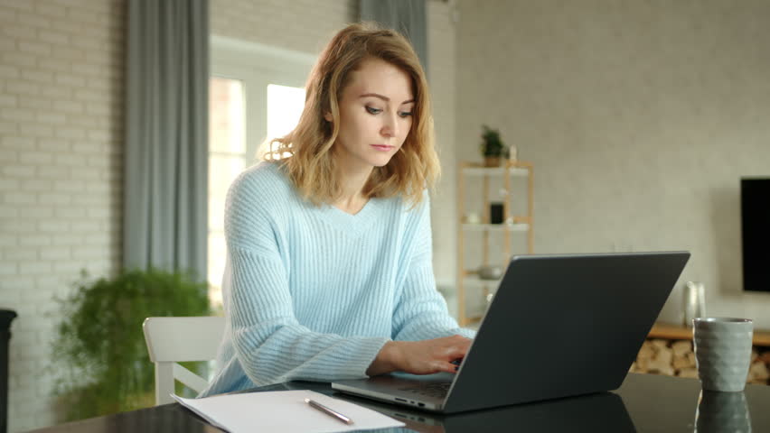 Young blonde is making notes on sheet of paper. The woman prints and searches for information in notebook. Beautifully furnished room in the background, High quality 4k footage