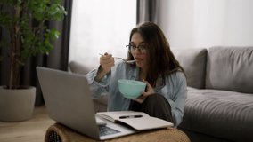 Woman working from home on laptop, making notes and eating cereal in between