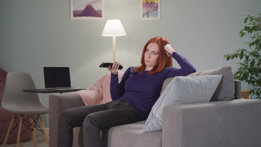 Powerless woman with ginger hair watches TV after long-time work in office. Housewife leans on hand using remote control to find interesting channel Royalty-Free Stock Footage #1102582567