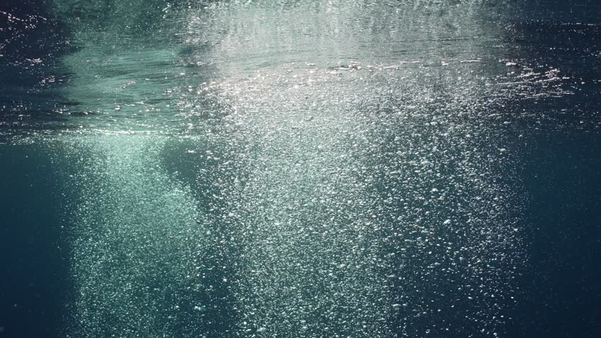 Motion of air bubbles rise up to surface in blue water in bright day sunlight, Slow motion. Sun beams penetrate water surface on bubbles rising underwater