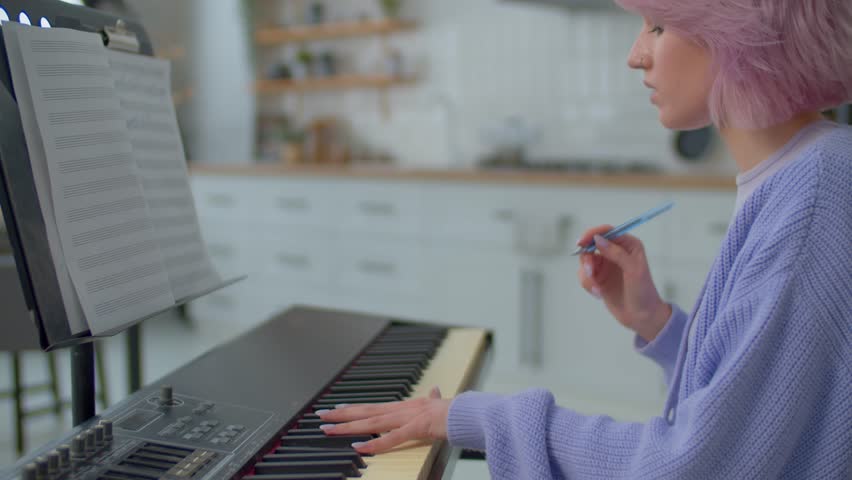 Close-up of inspired talented pretty pink haired female musician in creative process composing music using electronic keyboard, writing musical notes into lead sheet on sheet music stand. Side view.