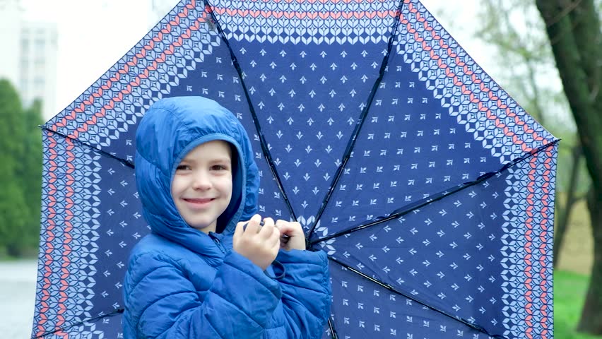 A cheerful 5-year-old boy stands under an umbrella, it is raining. Child in blue hooded jacket