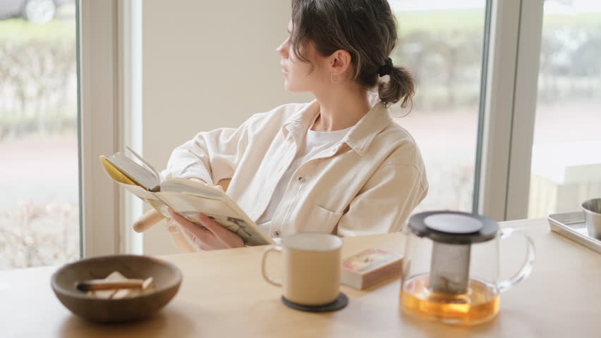 Young woman reading paper book sitting at table with mug of tea. Person spending time at home with her favorite hobbies, time to relax and slow down in cozy atmosphere. Stress relieving activities Royalty-Free Stock Footage #1102606413