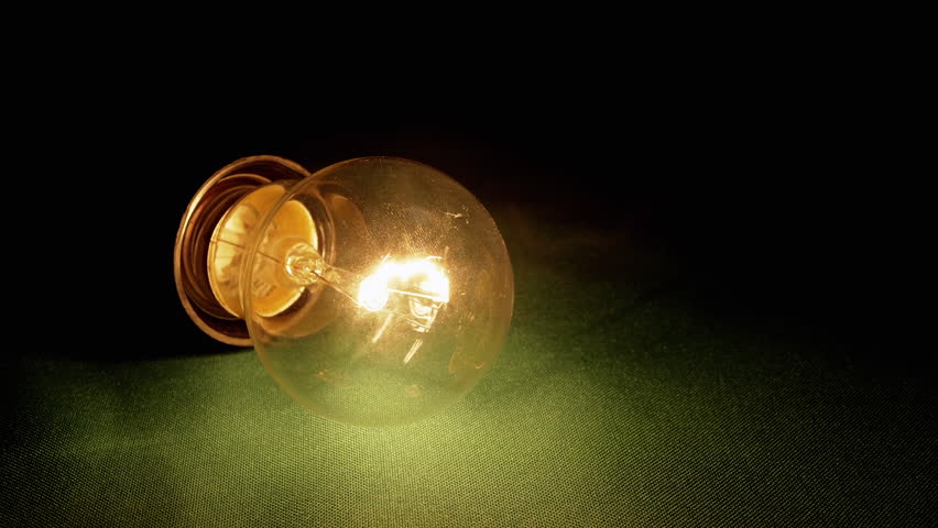 Yellow Incandescent Lamp Lights Up, Flickers on a Green Background in Dark Room. Close up. Tungsten light bulb with warm blinking light pulsates, lies on table. Power outage, power failure. Crisis. Royalty-Free Stock Footage #1102610621