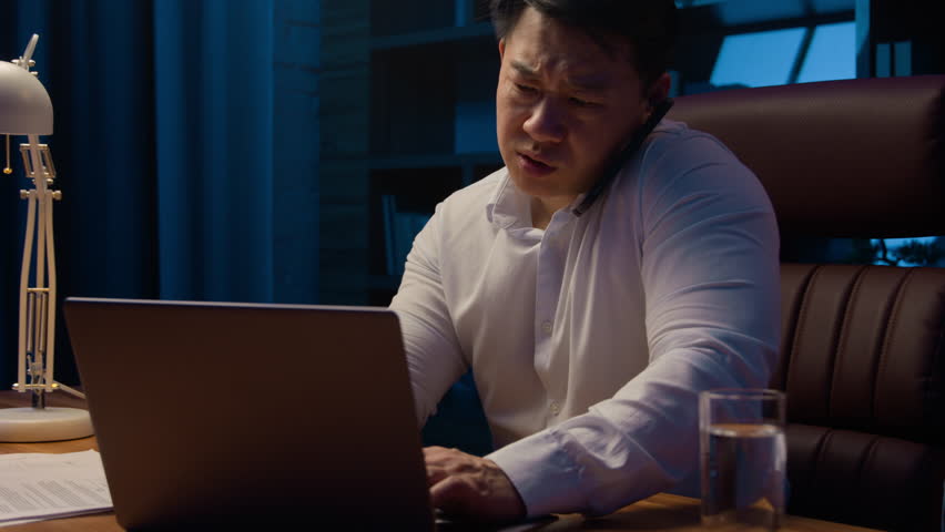 Tired angry Asian business man talking mobile phone at night evening office check documents working laptop overworked stressed Korean businessman executive manager arguing with call computer paperwork Royalty-Free Stock Footage #1102614143