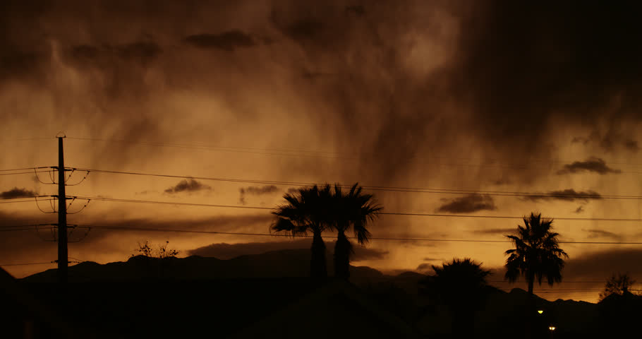 Apocalyptic dream-like vision with shafts of virga, heavy downpour, palm trees and transmission towers in silhouette against the menacing glow of a turbulent sky at dusk.