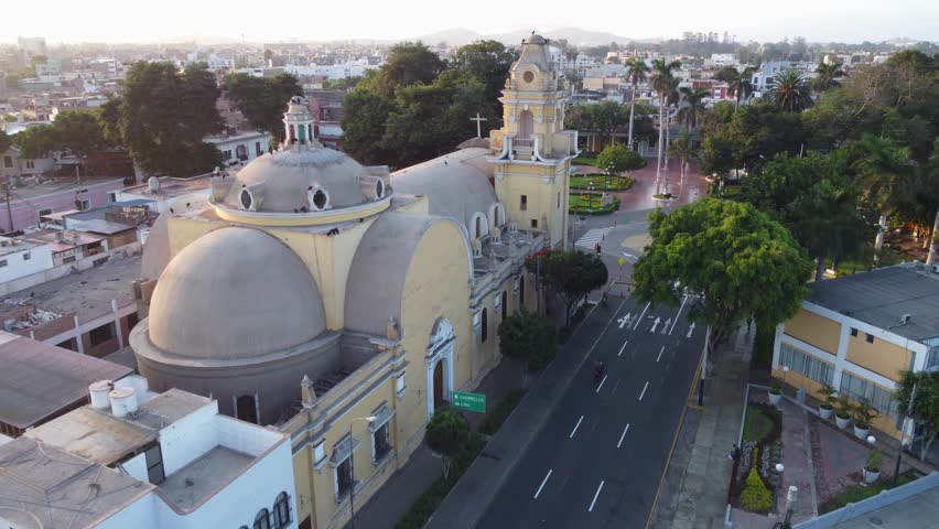 Drone flies over a church and focuses down on a park with many green trees. Recorded in early morning light. Located in "Parque Municipal de Barranco" in Lima, Peru.