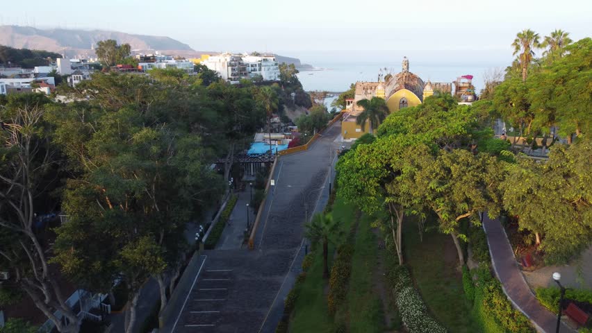 Drone flies above trees, below to the left is a small bridge called "Puente de los suspiros" to the right a church, in the background is the pacific ocean. Located in Barranco district of Lima, Peru.