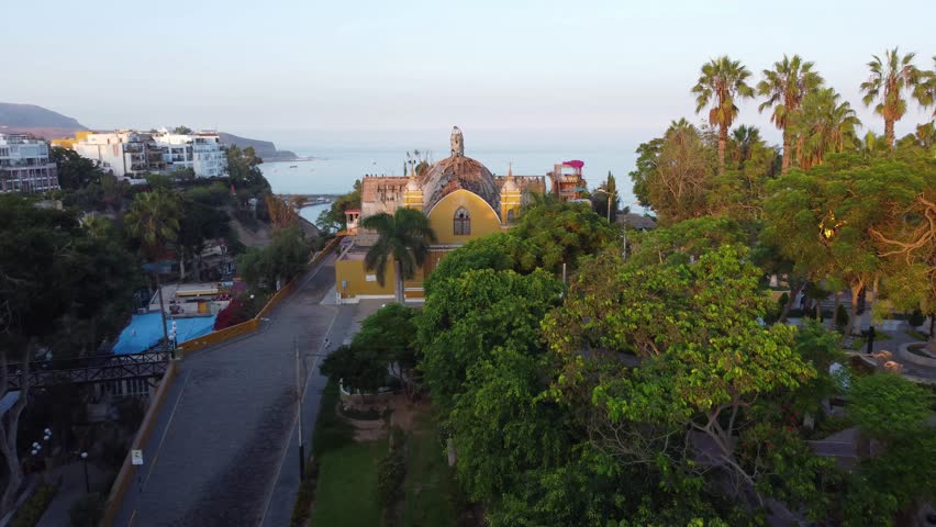 An old closed down church with a crumbling roof surrounded by green trees and with the pacific ocean behind it. Church is called "Iglesia La Ermita" Located in Barranco district of Lima, Peru.