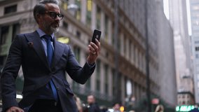 Adult Prosperous businessman in suit and stylish eye glasses stands on city street while calling on cellular. Smiling mature successful man talking on video mobile phone outdoors near office building