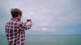 Man holds smartphone, takes photo or shoots video of arriving passenger airplane, airliner against the cloudy sky over the sea - back view, wide angle view. Photography, plane spotting concept