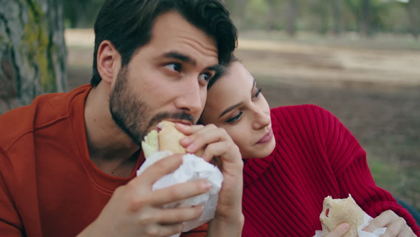 Couple relaxed tourists eating in forest enjoying beautiful nature view close up. Calm carefree family tasting sandwiches sitting outdoors. Attractive woman putting head on man shoulder feeling happy. | Shutterstock HD Video #1102616681