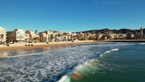 Panoramic drone aerial view of coastal city, old town of Sitges seen during sunny day. Close-up of surging Mediterranean sea waves. Province of Barcelona, Catalonia, Spain. Film intro. 4K video