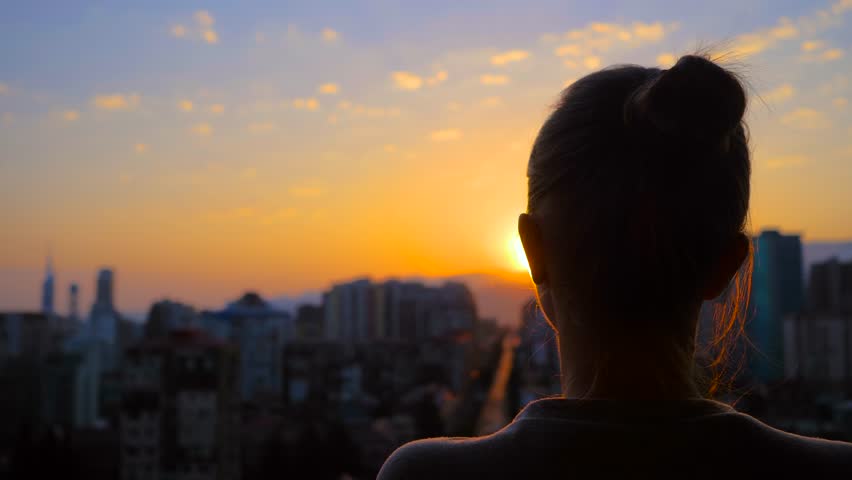 Back view: woman silhouette is standing on the balcony and looking at the sunset, sunrise sky over the city - close up, sun lens flares. Lonely, urban, dramatic and freedom concept Royalty-Free Stock Footage #1102618755