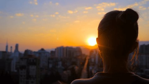 Стоковое видео: Back view: woman silhouette is standing on the balcony and looking at the sunset, sunrise sky over the city - close up, sun lens flares. Lonely, urban, dramatic and freedom concept
