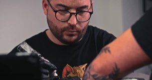 A professional tattooer artist doing picture on arm of man by machine black ink from a jar. Tattoo art on body. Equipment for making tattoo art. Master makes tattooed in light studio.