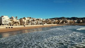 Panoramic drone aerial view of coastal city, old town of Sitges seen during sunny day. Close-up of surging Mediterranean sea waves. Province of Barcelona, Catalonia, Spain. Film intro. 4K video.