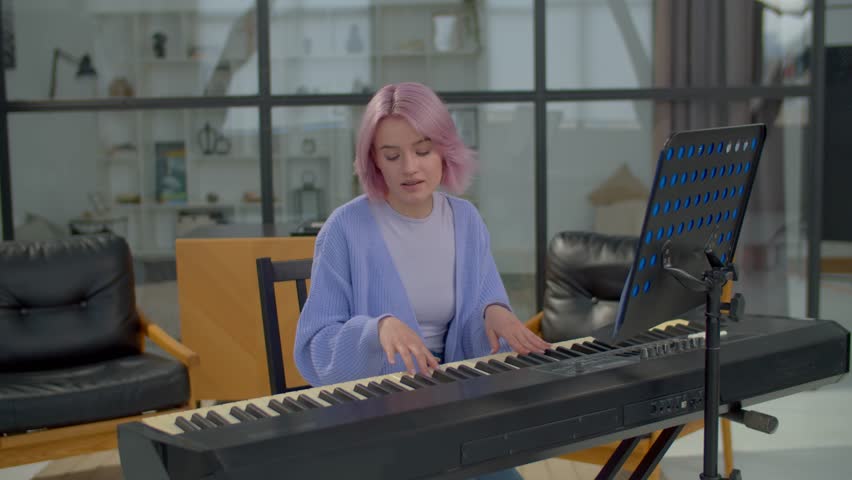 Talented attractive pink haired female songwriter composing melody for new song, playing piano keyboard and picking up chords, writing musical notes into music book on sheet music stand.