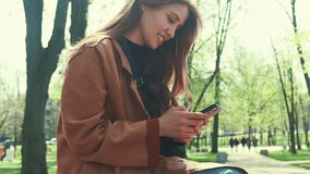Video of young smiling woman sitting on bench in city park and using smartphone during spring