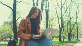 Young cheerful woman having online meeting on laptop while sitting outdoors in city