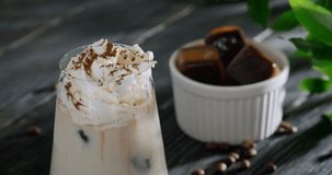 Iced coffee drink, latte with brown coffee ice cubes, milk and heavy cream topping, sprinkled with cinnamon powder, person putting a piece of chocolate on top, 4k close up footage, horizontal video