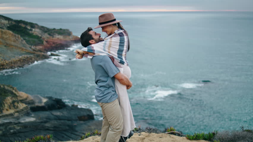 Loving young couple hugging on beautiful cloudy coast hill. Attractive bearded man lifting spinning smiling woman near calm ocean. Happy girl in hat kissing boyfriend at nature. Honeymoon love concept