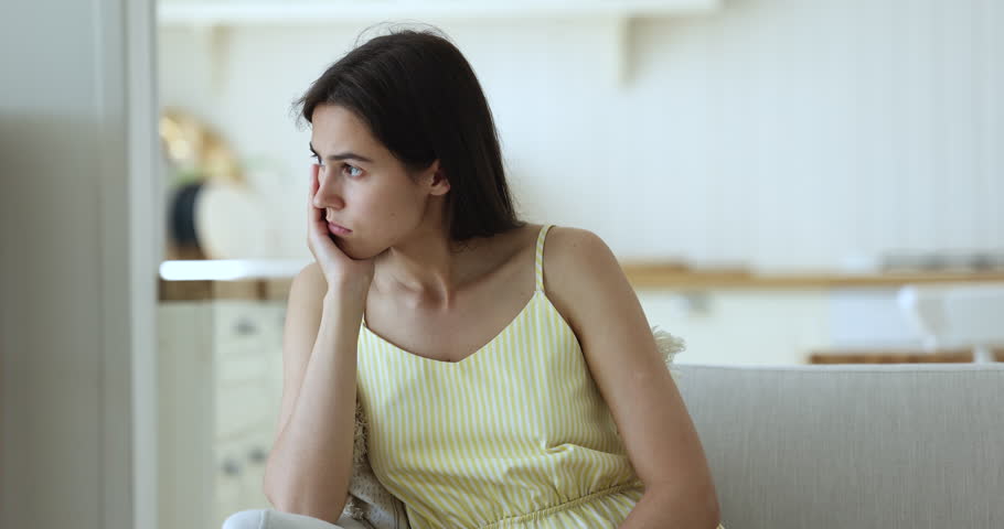 Pensive stressed young student woman sitting on sofa indoors, looking away, thinking over problems, concern, touching face, feeling tired, concerned, hurt, headache Royalty-Free Stock Footage #1102654961