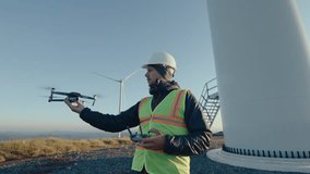 Engineer in uniform and hardhat launching a drone and using a controller when inspecting wind turbine at the wind farm. Medium long shot - Powered by Shutterstock - Get 15% off with code: PIKWIZARD15