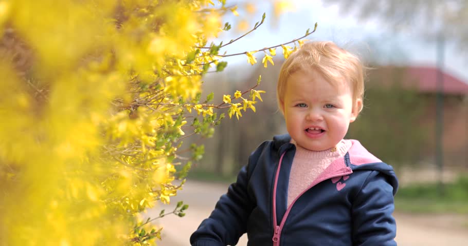 Beautiful baby girl playing with yellow flowers in nice spring sunny day outdoor. Blonde hair blue eyes todler. Slow motion shot 4K 120fps, high quality. Bouncing tree branches. Discovering new things