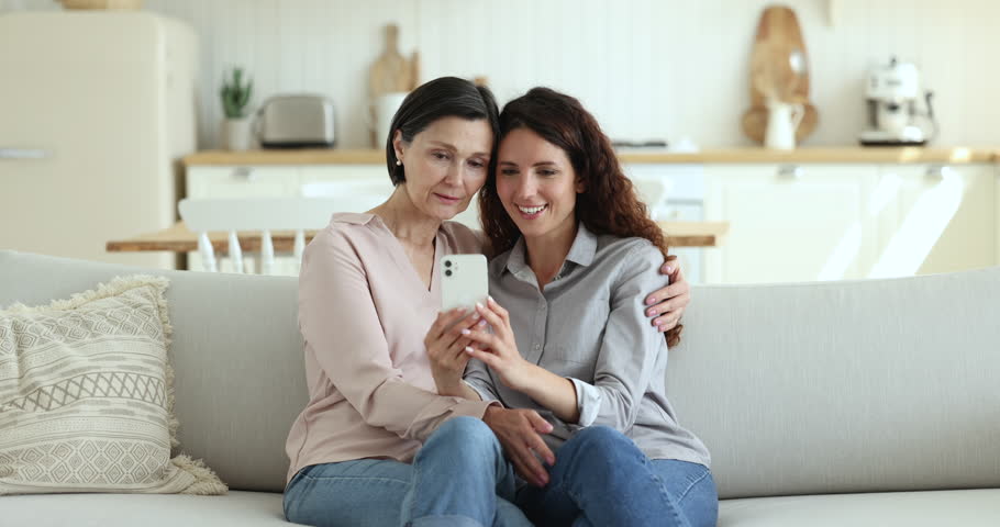 Happy pretty mature mother and beautiful adult daughter talking on video call on smartphone, taking selfie together, using mobile phone on home sofa, relaxing, enjoying leisure, online communication Royalty-Free Stock Footage #1102664633