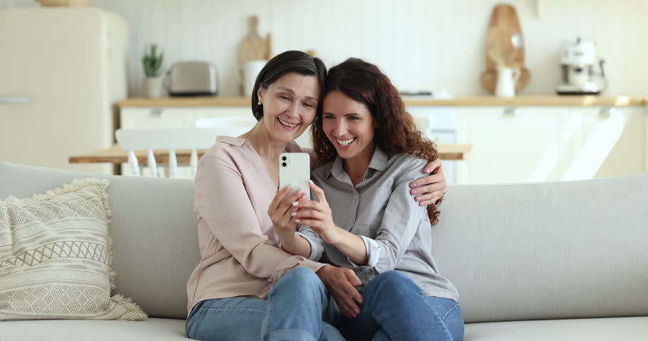 Happy pretty mature mother and beautiful adult daughter talking on video call on smartphone, taking selfie together, using mobile phone on home sofa, relaxing, enjoying leisure, online communication