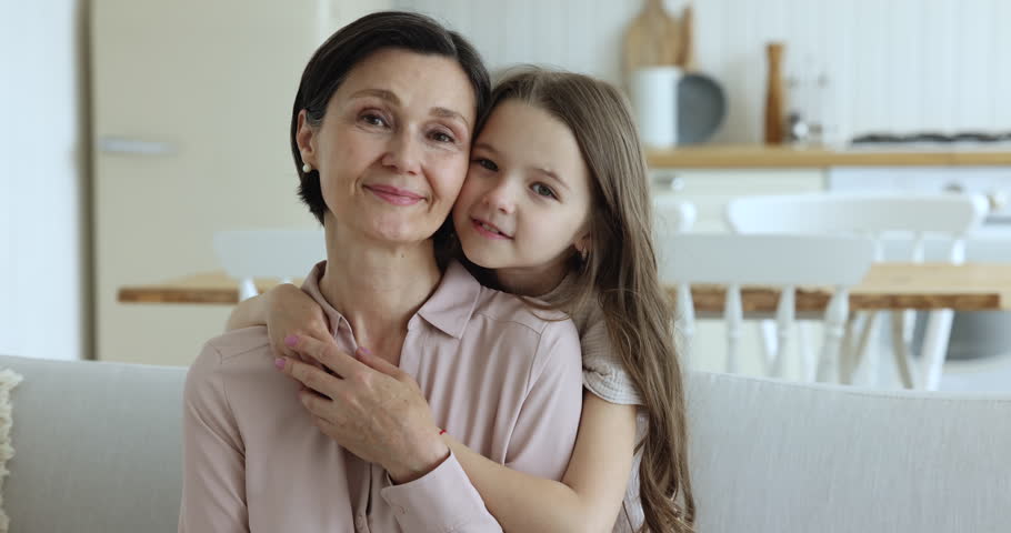 Mature 50s grandmother and cute grandkid child enjoying leisure together on home couch, looking at camera, laughing, feeling joy, cheerful, happy. Kid hugging grandma with love, affection Royalty-Free Stock Footage #1102664641