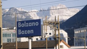 Bolzano train station sign with city name, train passing by, mountains on the background. Trentino, Italy