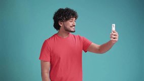 handsome cheerful smiling Indian man extends hand with smartphone, taking selfie portrait isolated over . Hispanic guy photographs himself, streaming live in social media in coral t-shirt on blue