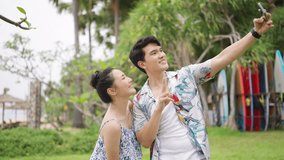 Happy Asian couple using mobile phone taking selfie together at seaside in sunny day. Man and woman enjoy and fun outdoor lifestyle travel at tropical island beach on summer holiday vacation.