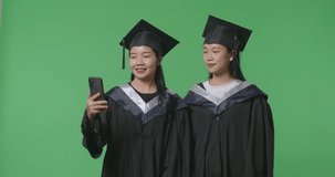 Asian Woman Students Graduate In Caps And Gowns With Diplomas Having A Video Call On Smartphone While Standing On The Green Screen Background In The Studio
