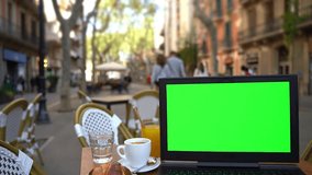 Laptop with green screen on the table in European street cafe. no hands typing. Static scene. Template video footage notebook with chromakey mock up. Morning coffee with croissant in Barcelona
