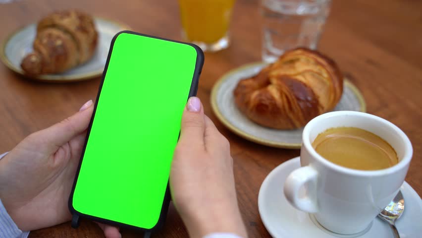 Mobile phone with green chromakey screen hand holding vertical portrait position. Hand swiping left and right. Street cafe breakfast with coffee  and croissant. Template video footage empty copyspace  Royalty-Free Stock Footage #1102668787