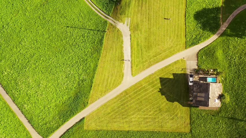 Aerial birds eye view of a luxurious two story swiss wooden chalet house with pool terrace and pick up car on green meadow gravel cross road, two people walking on grass field at sunset golden light