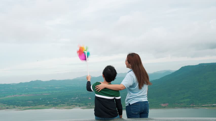 A progressive woman and her son are on vacation, enjoying the natural beauty of a lake at the bottom of a hill while the boy carries a toy windmill.