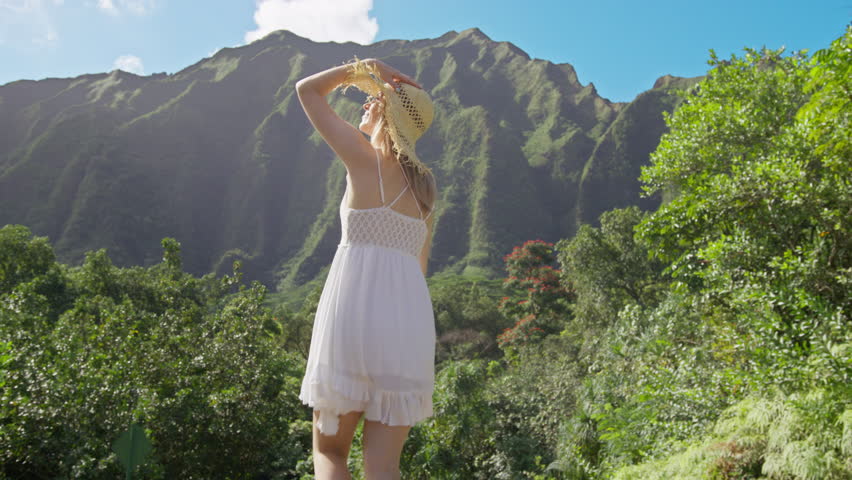 Stylish girl in straw hat and white boho dress with high jungle Hawaii mountain backdrop Oahu travel. Cheerful happy joyful woman tourist, happily raising hands up with huge green tropical plat leaf Royalty-Free Stock Footage #1102675439
