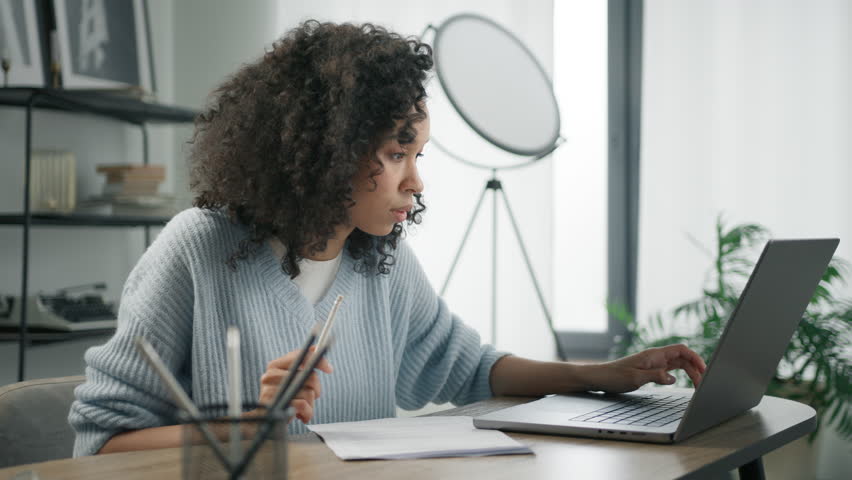 Focused and concentrated african female student e learning online on internet website. Young woman of color working remotely at home office desk, makes notes in work document, looks at laptop computer Royalty-Free Stock Footage #1102675493